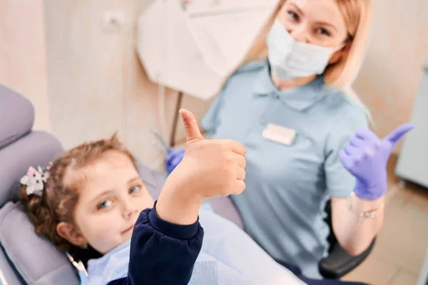女の子の手は親指をあげることに焦点を当てながら 医療マスクの女性歯科医は子供の横に座って 歯科処置後の承認ジェスチャーを示しています 小児科 歯科医療承認の概念 — ストック写真