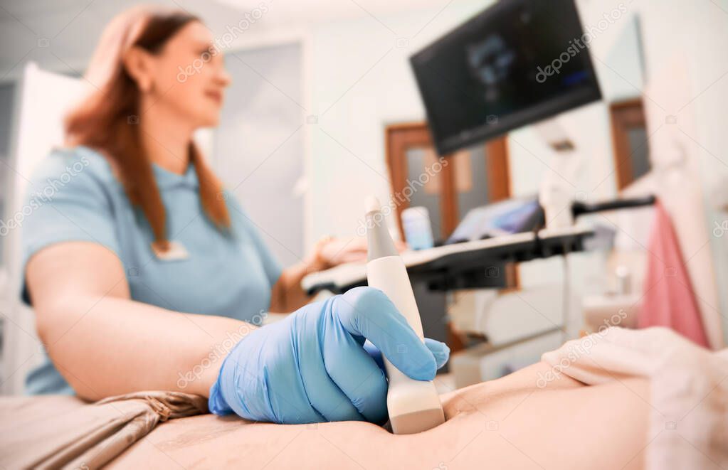 Focus on sonographer's hand in sterile glove examining woman with ultrasound scanner. Female doctor moving transducer on patient abdomen while looking at display. Concept of ultrasound diagnostics.