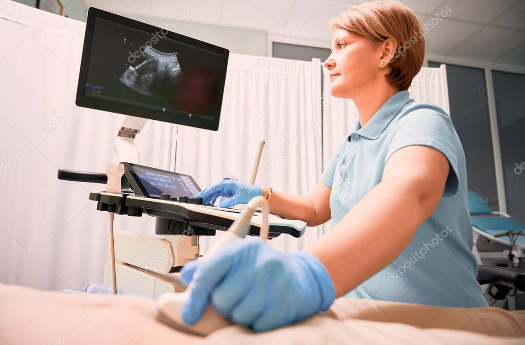 Female sonographer examining woman with ultrasound scanner. Doctor moving ultrasound transducer on patient abdomen while looking at display. Concept of medical examination and ultrasound diagnostics.