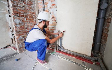 İş yerindeki sakallı genç adam daireye ısıtma boruları yerleştiriyor. Güvenlik kasklı bir erkek işçi su ve ısıtma sistemleri için bakım yapıyor. Tesisat ve tadilat kavramı.