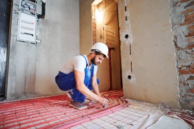 Mavi tulumlu ve beyaz miğferli genç tesisatçının yatay görüntüsü. Yeni apartman dairesinde yerden ısıtma sistemini kırmızı borularla bağlıyor. Kurulum çalışmaları kavramı