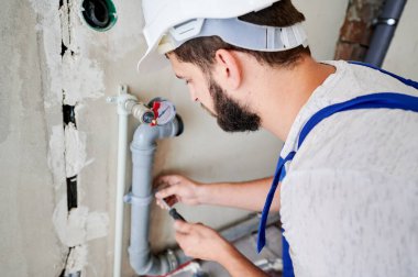 Gri kanalizasyon borularıyla çalışan genç sakallı tesisatçının yatay görüntüsü tornavidayla duvara tutturuluyor. Yakın plan. Bir yüze seçici odaklanma. Tesisat işi konsepti.