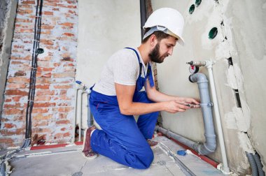 Gri kanalizasyon borularıyla çalışan bir tesisatçı tornavida yardımıyla onları duvara yapıştırıyor. Mavi üniformalı ve beyaz kasklı bir adamın dizlerinin üstünde duruşu. Tesisat işleri, ev yenileme konsepti.