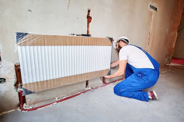 Male plumber in work overalls installing heating radiator in empty room. Young man in safety helmet installing heating system in apartment. Concept of installation, plumbing works and home renovation.