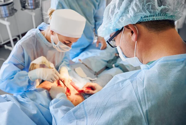 Medical workers in blue surgical suits performing plastic surgery in hospital. Doctor and assistant removing excess fat from patient abdomen. Concept of abdominoplasty and cosmetic surgery.