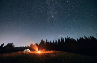 Siluet, erkek yürüyüşçünün yıldızlı güzel gece gökyüzünün altında kamp ateşinin yanında durduğunu gösteriyor. Çimenli tepenin altında çadırlı mavi yıldızlı gökyüzü manzarası. Seyahat, yürüyüş, gece kampı kavramı.