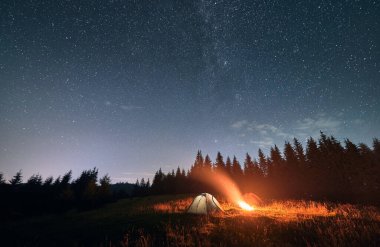 Yıldızlı gökyüzünün altında gece kampı. Dağlardaki güzel manzaranın geniş açılı görüntüsü. Arka planda iki turist çadırı ve kamp ateşi var. Seyahat ve astrofotoğrafçılık kavramı