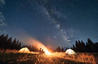 Kamp çadırları ve kamp ateşinin yakınındaki yürüyüşçülerle dağlık vadideki yıldızlı gökyüzünün muhteşem manzarası. Genç kadın ve erkek yolcular şenlik ateşinin yanında oturuyorlar. Gece kampı ve galaksi kavramı.