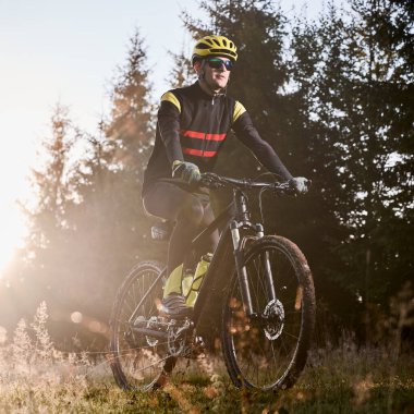 Bisikletli genç adam çimenli tepede bisiklet sürüyor. Sabah güneşi ve arka planda ağaçlar var. Dışarıda bisiklet sürerken güvenlik kaskı ve gözlük takan erkek bisikletçi. Kare resim