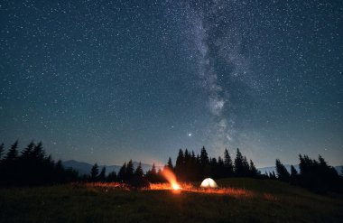 Dağlardaki güzel manzaranın geniş açılı görüntüsü. Güzel yıldızlı gökyüzünün altında turist çadırları ve kamp ateşi. Dağlar çayırların arkasında. Gece kampı ve astrofotoğrafçılığı kavramı