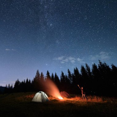 Kamp ateşinin yanında yürüyen bir adam ve yıldızlı bir gece gökyüzünü işaret ediyor. Gezgin, şenlik ateşi ve kamp çadırlarıyla dağlık vadinin altında mavi yıldızlı gökyüzü manzarası. Yürüyüş kavramı, gece kampı..