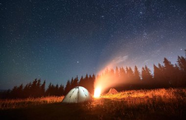 Yıldızlı gökyüzünün altında gece kampı. Dağlardaki manzara çok güzel. İki aydınlatılmış turist çadırı ve kamp ateşi, arka planda çelenk duvarı. Seyahat ve astrofotoğrafçılık kavramı