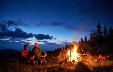 Ateşin başında oturan iki yürüyüşçü ve dağlarda sıcak bir akşamın tadını çıkarıyorlar. Birkaç arkadaş kamp yapıyor ve alacakaranlıkta kamp ateşi yakıyor..