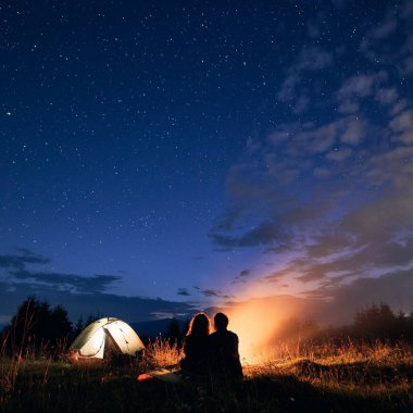 Güzel bir gece manzarası çimenli tepenin üzerinde parlayan kamp çadırı, kamp ateşi ve yürüyüşçülerle. Birkaç turistin şenlik ateşinin yanında dinlendiklerini gördüm. Gece kampı kavramı, ilişki..
