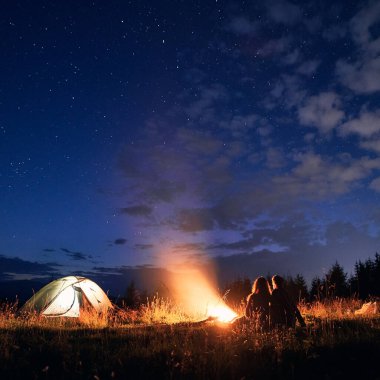 Güzel bir gece manzarası çimenli tepenin üzerinde parlayan kamp çadırı, kamp ateşi ve yürüyüşçülerle. Şenlik ateşinin yanında dinlenen kadın ve erkek turistlerin silueti. Gece kampı kavramı, ilişki..