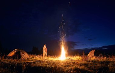 Kamp çadırları, kamp ateşi ve yürüyüşçülerle çimenli tepenin üzerinde bulutlu bir gece. Genç adam ve kadın gezginler mavi gece gökyüzünün tadını çıkarıyorlar. İlişki kavramı, gece kampı ve seyahat..