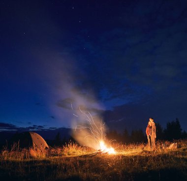 Kamp çadırları, kamp ateşi ve yürüyüşçülerle çimenli tepenin üzerinde gece yıldızlı gökyüzü. Genç adam ve kadın gezginler mavi gece gökyüzünün tadını çıkarıyorlar. İlişki kavramı, gece kampı ve seyahat..