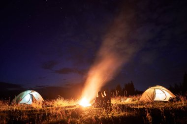Birkaç turistin birlikte kamp yaparken çekilmiş yatay görüntüsü. Genç bir adam ve bir kadın, doğada akşam vakti kamp ateşinin yanında oturuyorlar. Yanlarda parlayan çadırlar