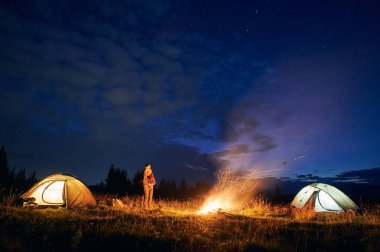 Birkaç turistin birlikte kamp yaparken çekilmiş yatay görüntüsü. Genç bir oğlan ve kız, doğada akşam vakti kamp ateşinin yanında dururlar. Yanlarda parlayan çadırlar