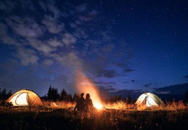 Gece manzarası, çimenli tepenin üstündeki yıldızlı gökyüzü. Kamp çadırları, kamp ateşi ve yürüyüşçülerle dolu. Genç turistler şenlik ateşinin yanında dinleniyorlar. Yürüyüş, gece kampı ve ilişki kavramı.