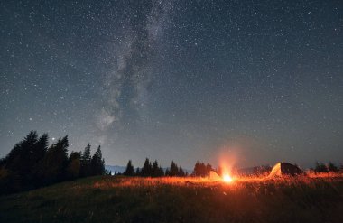 Samanyolu 'yla muhteşem yıldızlı gökyüzünün altında gece kampı. Dağlardaki manzara çok güzel. Arka planda iki çadır ve kamp ateşi var. Seyahat ve astrofotoğrafçılık kavramı