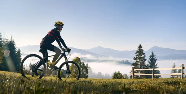 男人骑自行车在长满青草的山上 望着美丽的薄雾的群山 骑自行车的男性骑自行车者欣赏高山壮丽的全景 自行车和自然的概念 — 图库照片