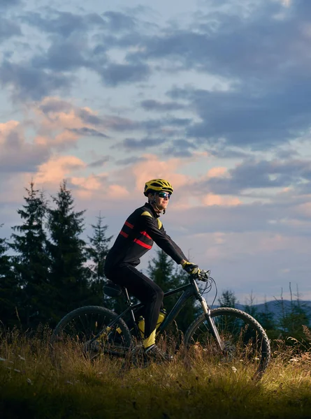 年轻人骑自行车在日落时在山上骑自行车 骑自行车的人戴着头盔 戴着运动眼镜 身穿制服 侧视图 背景是夜空 积极生活方式的概念 — 图库照片