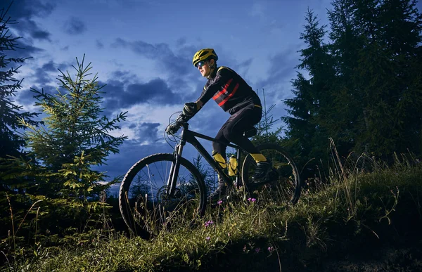 年轻男子骑自行车下山 背景是美丽的蓝色夜空 骑自行车的男性骑自行车者在夜间骑着自行车沿着青草丘陵地带骑行 骑单车和积极休闲的概念 — 图库照片