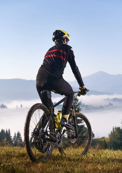 Back view of young man in cycling suit riding bicycle on grassy hill. Male bicyclist enjoying the view of majestic mountains during bicycle ride. Concept of sport and bicycling. Vertical picture.