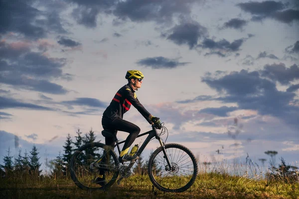 年轻人骑自行车在日落时在山上骑自行车 骑自行车的人戴着头盔 戴着运动眼镜 身穿制服 侧视图 美丽的夜空背景 积极生活方式的概念 — 图库照片