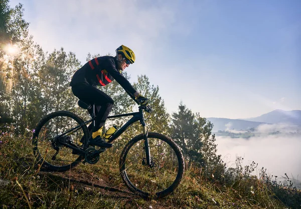 在雾蒙蒙的清晨 年轻人骑着自行车在山上下山 太阳正透过树冠闪耀 背景上的云彩达到顶峰 积极生活方式的概念 — 图库照片
