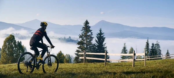 男人骑自行车在长满青草的山上 望着美丽的薄雾的群山 骑自行车的男性骑自行车者欣赏高山壮丽的全景 自行车和自然的概念 — 图库照片