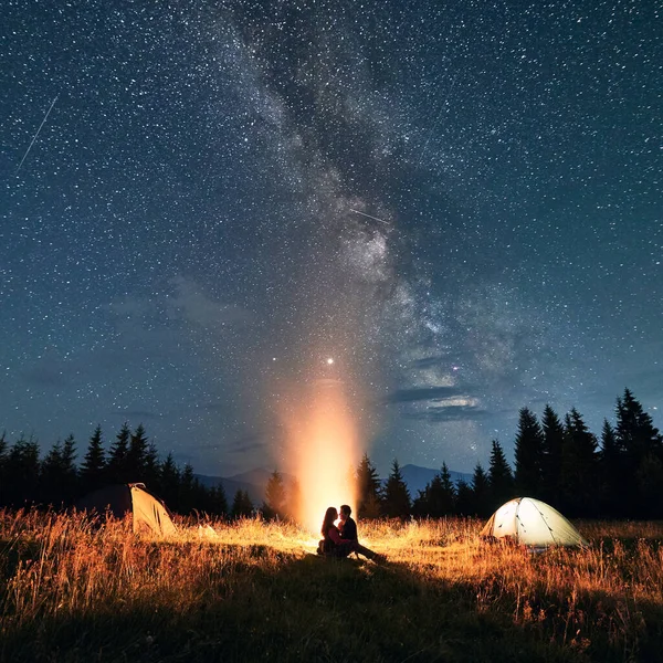 View of night starry sky over mountain valley with camp tents and hikers near campfire. Young woman and man travelers sitting under magical sky with stars. Concept of night camping and relationship.