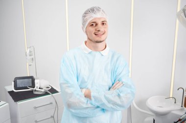 Tıp şapkası, palto giyen, beyaz ofiste kolları çapraz duran neşeli genç doktor asistanının portresi. Kameraya bakıp gülümsüyor. Diş hekimi ve tıp konsepti.