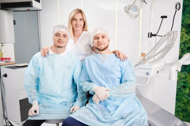 Tıbbi koruyucu giysiler içinde yakışıklı erkek stomatologlar kameraya bakıp gülümserken çekici kadın doktor erkeklerin arkasında duruyor. Diş hekimliği ve diş hekimi kavramı.