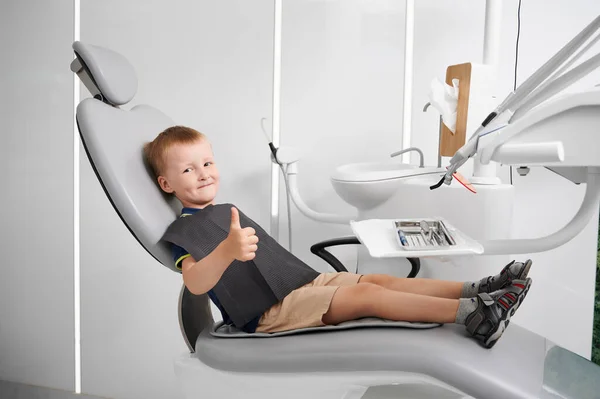 Little boy wearing black bib, sitting in dentist chair, showing thumb up. Well equipped dentist office. Concept of pediatric stomatology, dentistry and dental care.