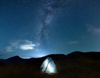Gecenin panoramik manzarası çimenli tepenin üzerinde parlayan turist çadırıyla yıldızlı gökyüzü. Dağ vadisi manzarası. Mavi gökyüzünün altında kamp çadırı var. Yıldızlar ve Samanyolu. Gece kampı kavramı.
