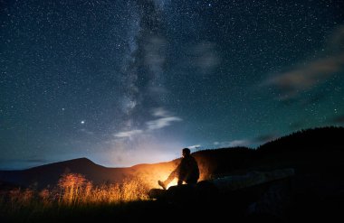 Ormanda rahatlayan ve ateş yakan bir adamın silueti. Erkek turist, yıldızlı gökyüzü ve Samanyolu 'nun altındaki dağlarda şenlik ateşinin yanında oturuyor. Güzel manzara kavramı.