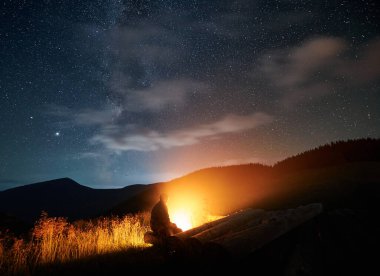 Turist doğada dinleniyor ve yıldızlı gökyüzünün altında şenlik ateşinin tadını çıkarıyor. Temiz havada rahatlama ve yıldızlarla gece gökyüzüne bakma kavramı..