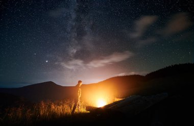 Dağlarda yürüyen ve ateş yakan bir adamın siluetinin yan görüntüsü. Erkek turist akşamları ormanda şenlik ateşinin yanında dinleniyor. Sağlıklı yaşam tarzı kavramı.