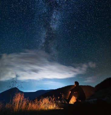 Ormanın yakınında rahatlayan ve gece gökyüzünü yıldızlarla ve Samanyolu 'yla izleyen insan siluetinin yan görüntüsü. Erkek turist akşam vakti dağlarda şenlik ateşinin yanında oturuyor. Seyahat kavramı.