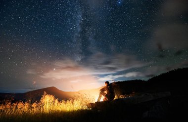 Şenlik ateşinin yanında oturup yıldızlara ve Samanyolu 'na bakan genç adamın yan görüntüsü. Güzel yıldızlı gökyüzü ile temiz havada dinlenme kavramı.