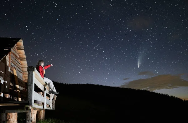 Side view of young man sitting and looking starry sky at home from balcony, pointing at comet Neowise. Concept of enjoying incredible nature and beautiful stars on sky at night.