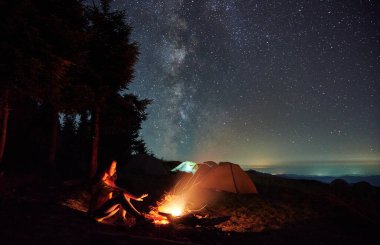 Güzel bir gece manzarası çimenli tepenin üzerindeki yıldızlı gökyüzü. Kamp çadırları ve kamp ateşi yakınında bir kız gezgin. Genç kadın yürüyüşçü, yıldızlı sihirli gökyüzünün altında şenlik ateşinin yanında elini ısıtıyor..