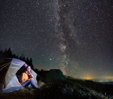 Kamp çadırları ve kadın gezginlerle çimenli tepenin üzerindeki yıldızlı gece manzarası muhteşem. Büyüleyici genç bir kadın turist çadırında yıldızlarla ve Samanyolu 'yla büyülü gökyüzünün altında oturuyor..
