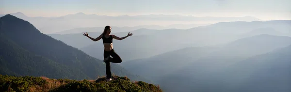 山と空を背景に芝生の丘の上でヨガポーズを行う若い女性のフィット感のパノラマビュー アウトドアでヨガを練習するスポーツウェアの美しい女性 スペースのコピー — ストック写真