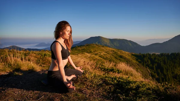 蓮の花の位置に芝生の丘の上に座ってスポーツウェアで魅力的な若い女性と屋外で瞑想しながらガイン泥手ジェスチャーを行います 山の中で瞑想ヨガの演習を行うスポーティ女性 — ストック写真