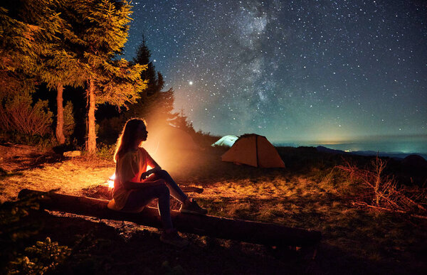 Side view of silhouette of woman relaxing near forest in campsite. Female sitting near bonfire and admiring starry sky. Concept of adventure and night camping in the mountains.