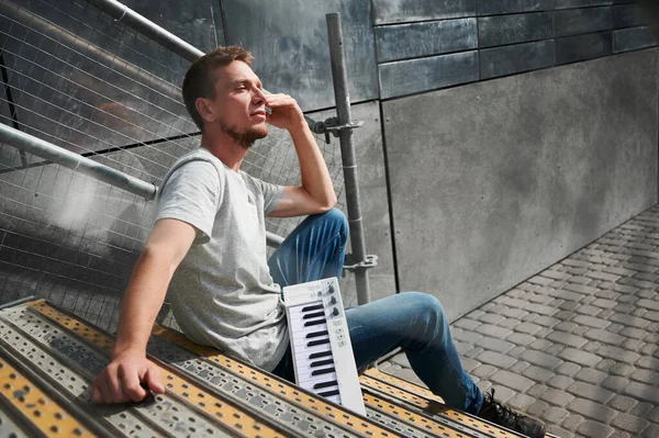 Side view of male with piano keyboard is sitting on stairs. Young man in grey T-shirt is resting on stairs on street and enjoy sunny weather. Concept of free time.