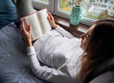 Pencere kenarına uzanmış kitap okuyan alımlı genç bayanı yakından çek. Beyaz kazaklı güzel bir kadın evde dinlenirken ilginç romanların tadını çıkarıyor. Okuma, edebiyat ve boş zaman kavramı.